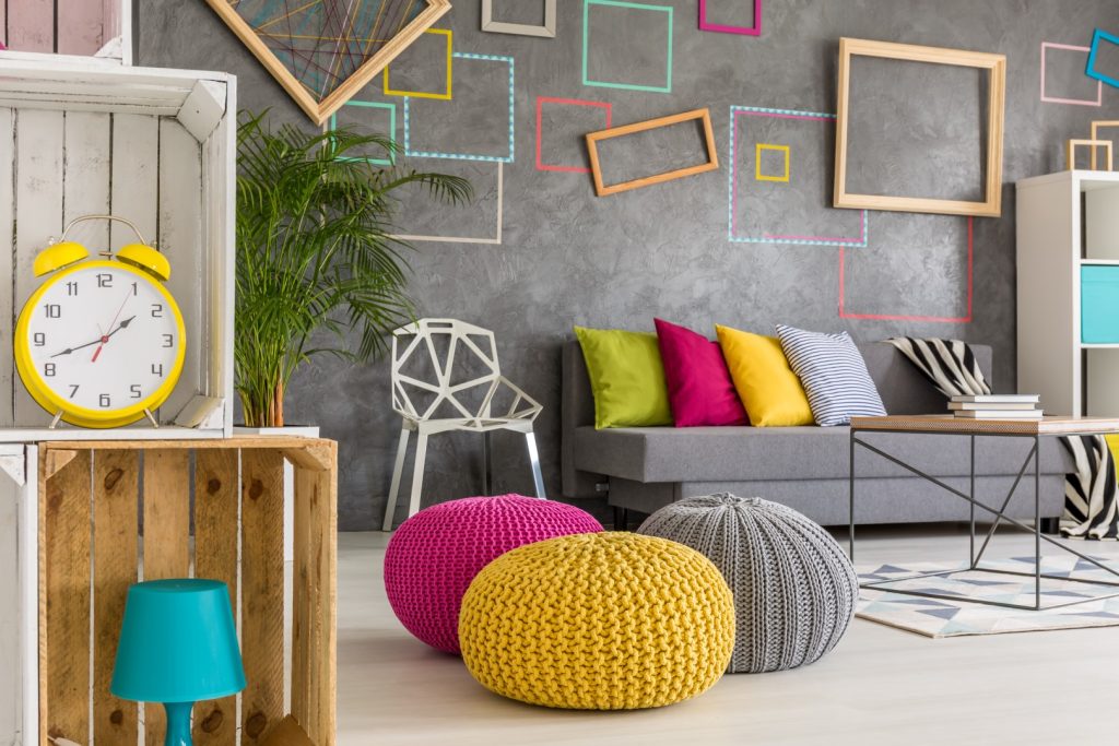 A colorful modern living room features a gray sofa, bright cushions, knitted poufs, a yellow clock, and playful geometric wall art, creating a cozy, cheerful mood.