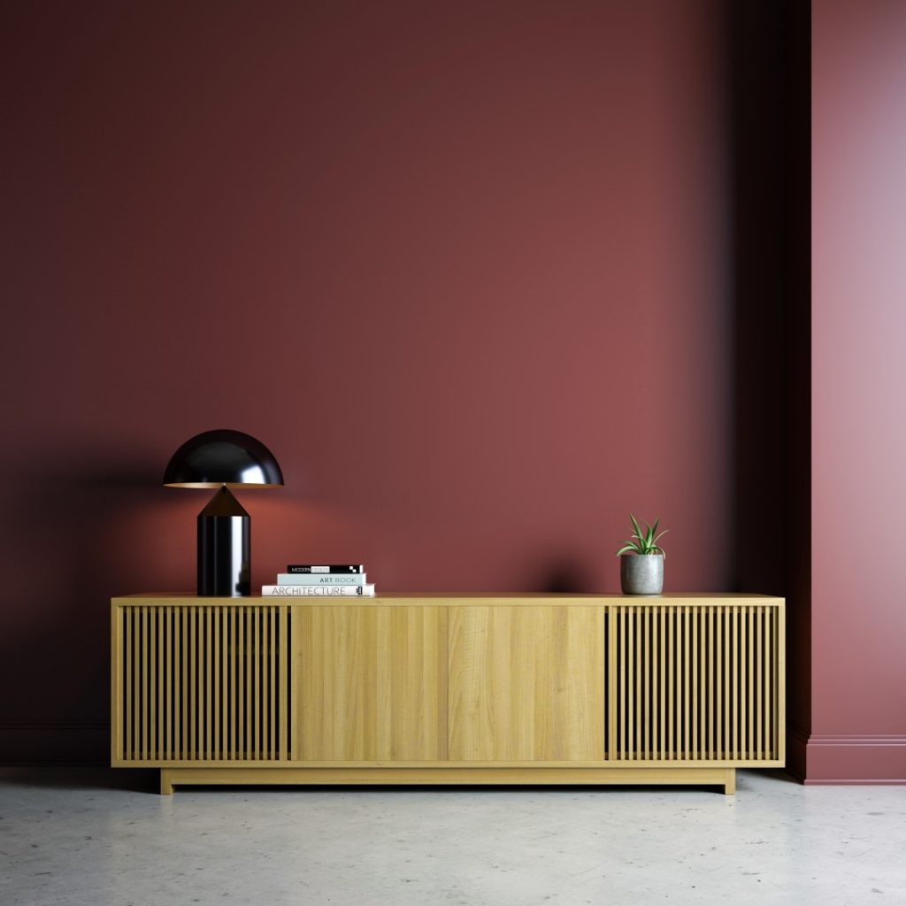 A minimalist mid-century modern wooden sideboard with slatted doors stands against a muted rose wall, topped by a black lamp, books, and a small plant.