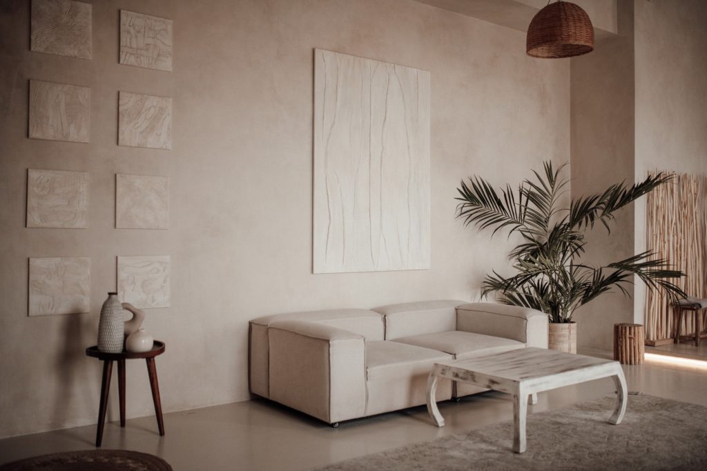 A serene beige living room with a cream sofa, sculptural coffee table, framed abstract art, leafy palm, and woven pendant lamp creates a calm, minimalist mood.