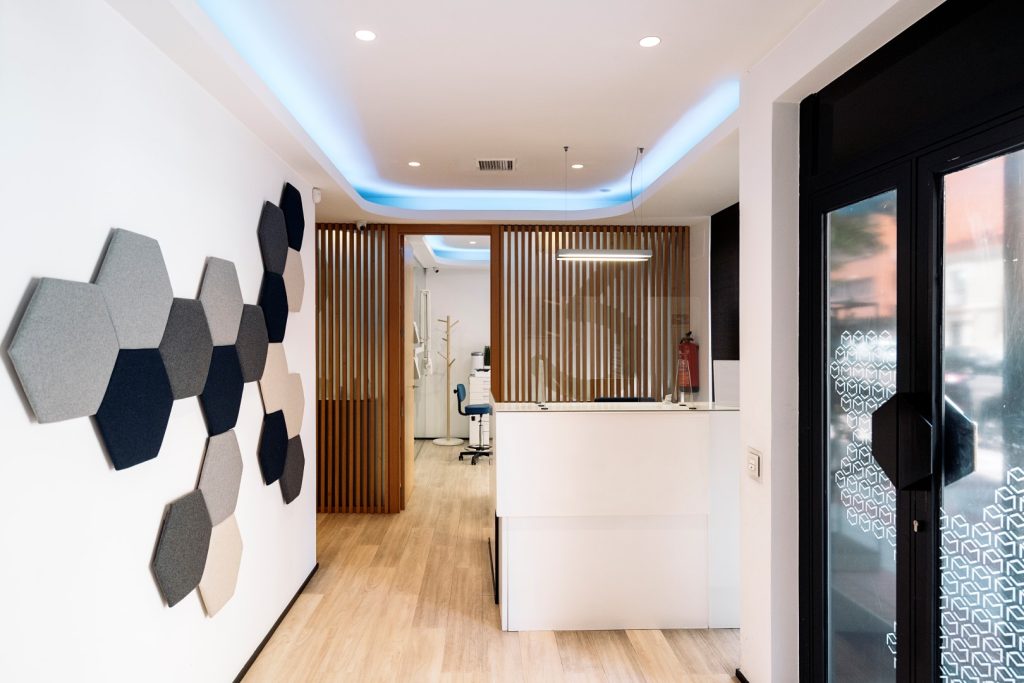 Bright, modern clinic reception with a white desk, wood-slat partition, hexagonal wall panels, and glass entrance; clean, calm, professional feel.