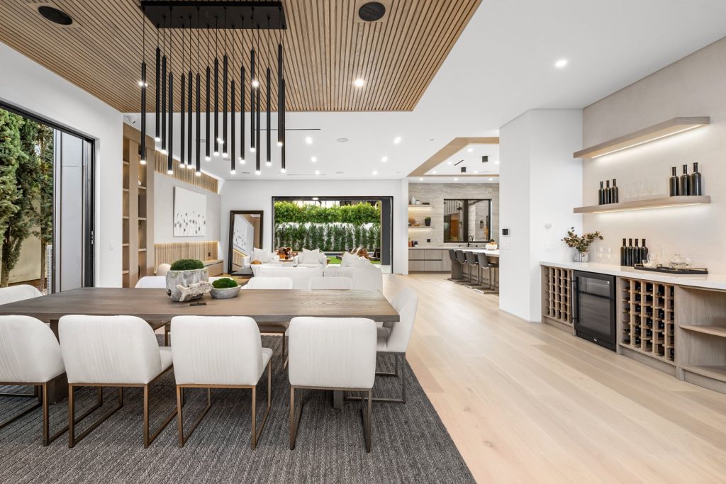 Bright modern open-plan dining and kitchen with a long wood table, upholstered chairs, wine storage, and garden views, creating an airy, elegant feel.