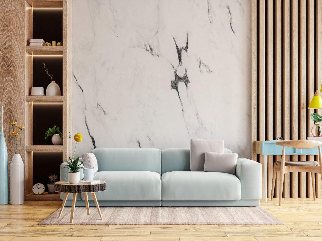 Calm modern living room with a pale blue sofa beneath dramatic veined marble, warm wood shelves and slats, plants, and a small round table.