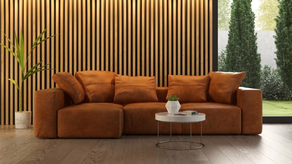 Caramel-brown sectional sofa and round coffee table sit before a wood-slat wall; a potted plant and garden view create a calm, modern living room.