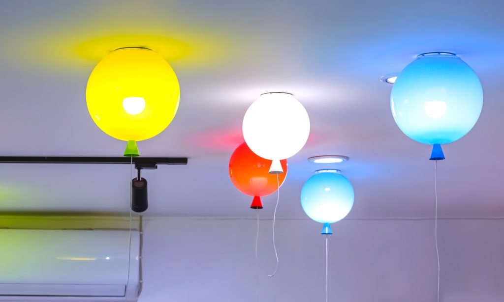 Five glowing balloon-shaped ceiling lights—yellow, white, red, and blue—hang above a room, casting colorful halos and creating a playful, festive mood.