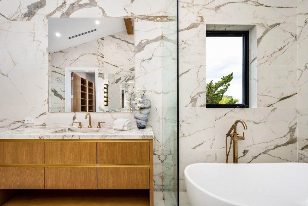 Luxurious marble bathroom with a wood-front vanity, brass faucets, white bathtub, and black-framed window overlooking greenery; serene and elegant.