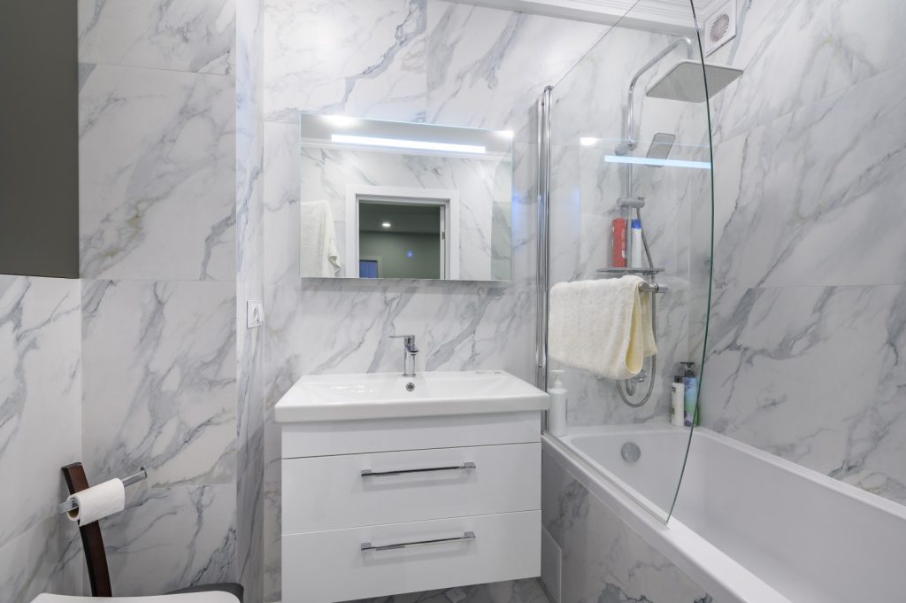 Modern bathroom with white marble-patterned walls, a vanity and mirror, and a bathtub enclosed by glass; the clean, bright space feels calm and polished.