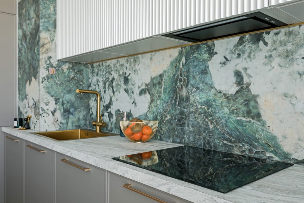 Modern kitchen with a dramatic green-and-white marble backsplash, brass faucet, bowl of oranges, and black glass cooktop; refined, calm luxury.