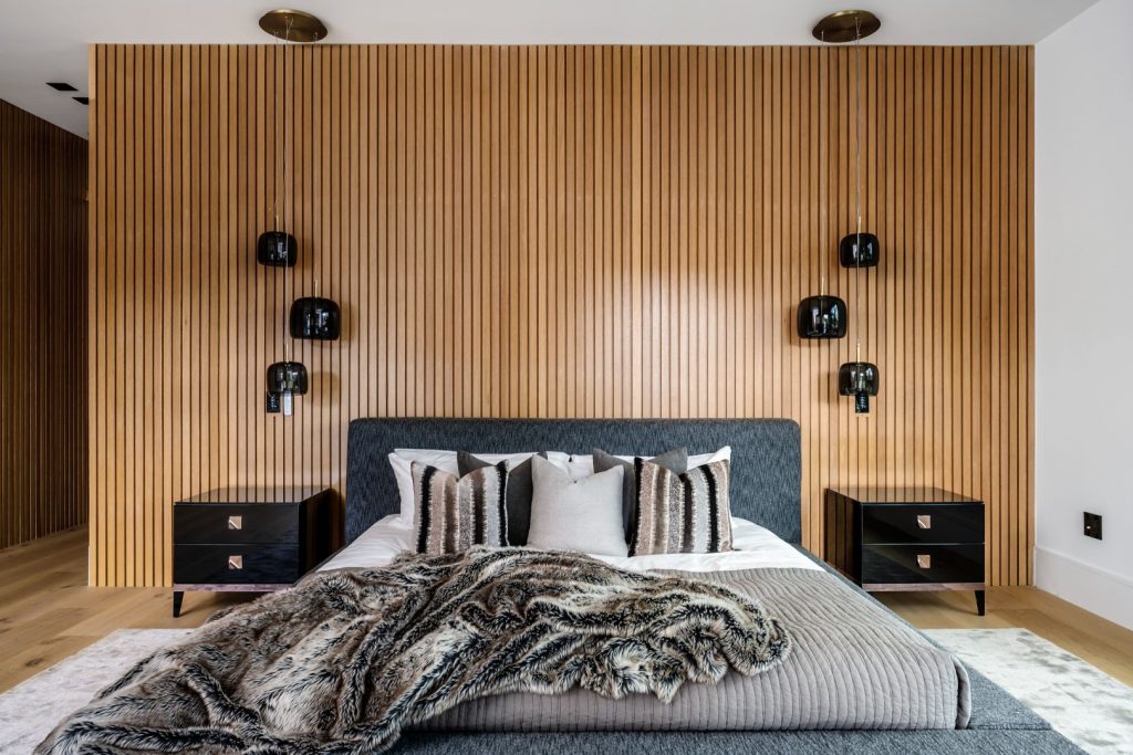 A serene modern bedroom with a king bed, layered neutral bedding and a dark fur throw, framed by a warm vertical wood-slat wall and black sconces.