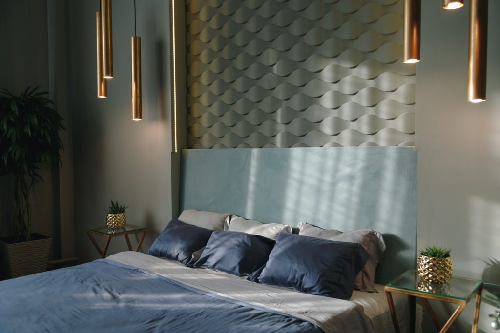 A serene modern bedroom with a blue bed, layered pillows, sculpted wall panels, warm brass pendant lights, and leafy plants in soft daylight.