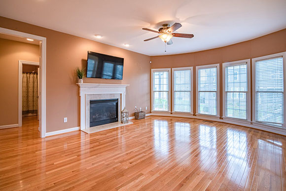 Spacious living room with hardwood floors, beige walls, a ceiling fan, a wall-mounted TV above a white fireplace, and large bay windows with blinds.