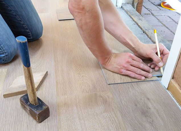 Person kneeling and marking measurements on wood flooring near a door frame, with a hammer placed on the floor beside them.