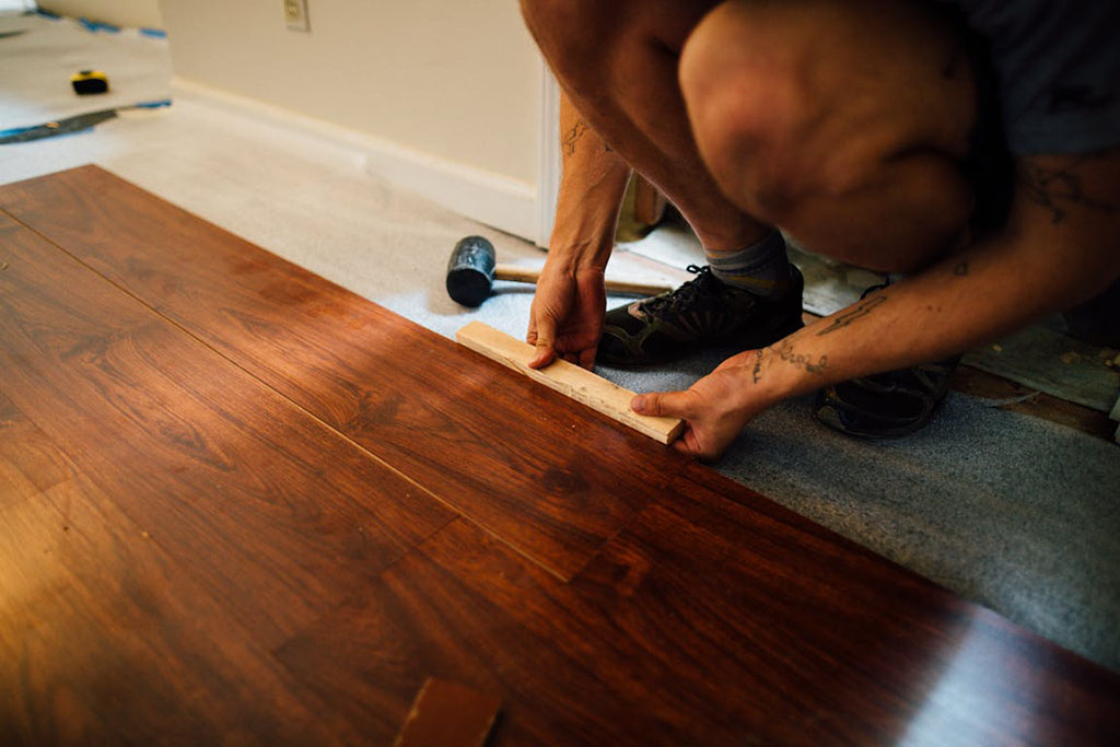 Person installing dark wood laminate flooring using a wooden block and rubber mallet, kneeling on a gray underlayment.