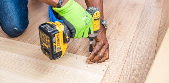 Person wearing green gloves using a yellow cordless drill to screw into wooden flooring or paneling.