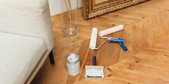 Paint rollers, a paint can, and a brush on a wooden floor next to a beige sofa and a large framed mirror.