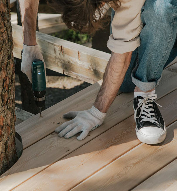 A person wearing gloves and sneakers is using a power drill to secure wooden boards on an outdoor structure.